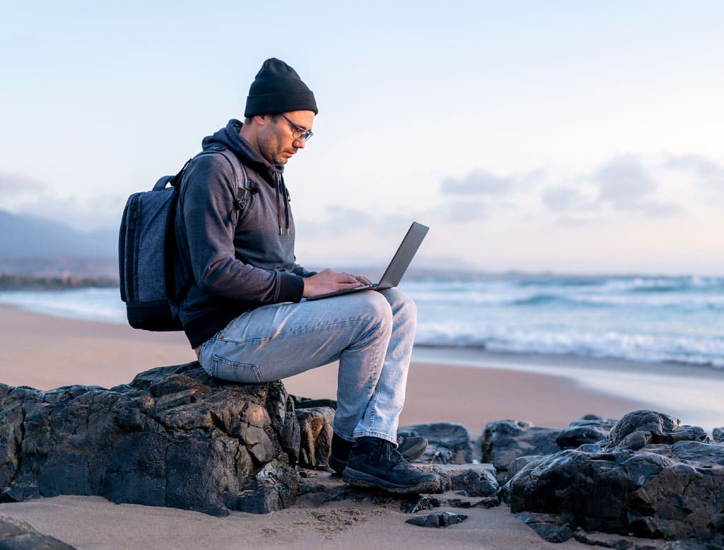 Un hombre sentado sobre un piedra en una playa consulta su portátil